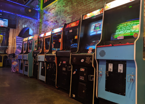 A row of vintage arcade machines in a dimly lit space with colorful lights and exposed brick walls.