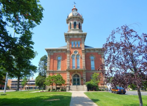 Historic brick building with a clock tower, surrounded by trees and greenery on a sunny day.