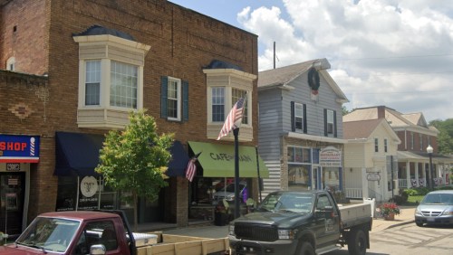 A quaint town street featuring brick buildings, shops, and American flags under a partly cloudy sky.