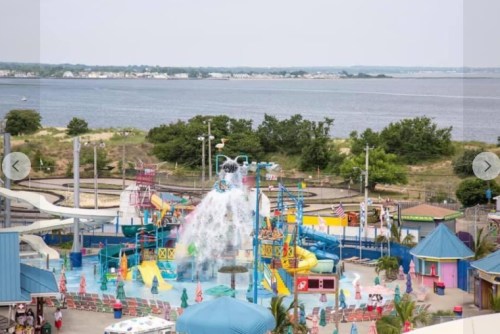 A colorful water park with slides and a splash area, overlooking a calm body of water and green trees in the background.