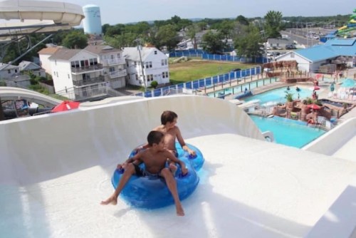 Two boys sit on a blue inner tube at the top of a water slide, ready to descend into a pool below.