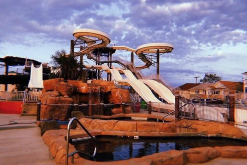 Water slides at a pool area, surrounded by rocks and palm trees, under a cloudy sky.