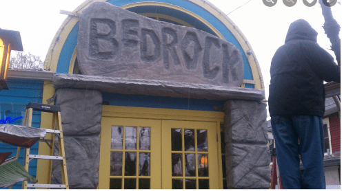 Sign reading "BEDROCK" on a stone archway above a yellow door, with a person in a hooded jacket nearby.