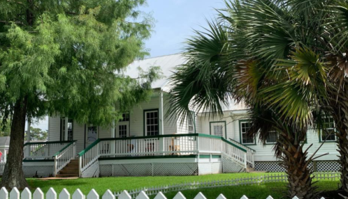 A white house with a green porch, surrounded by palm trees and a white picket fence, under a clear blue sky.