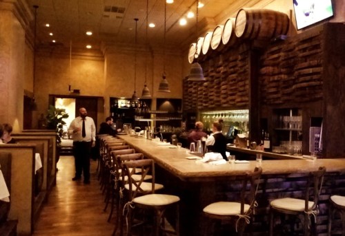 A cozy restaurant interior featuring a wooden bar, barrels, and patrons dining at tables. A waiter stands nearby.