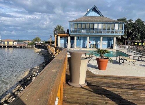 A wooden boardwalk leads to a blue building by the water, with a pool and lounge chairs under a cloudy sky.