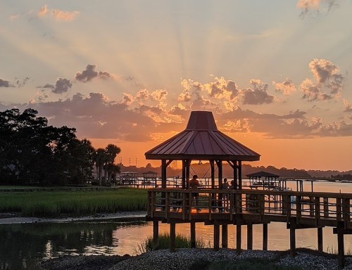 A gazebo by the water at sunset, with rays of light shining through clouds and silhouettes of people inside.