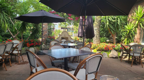A serene outdoor dining area with empty tables, umbrellas, and lush greenery in a garden setting.