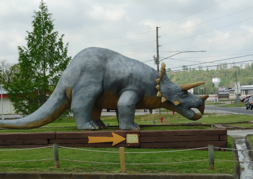 A large dinosaur statue, resembling a triceratops, stands on a wooden base with a directional sign nearby.