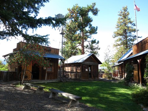 A rustic outdoor scene featuring wooden buildings, a windmill, and tall trees, with benches and a flag in the background.