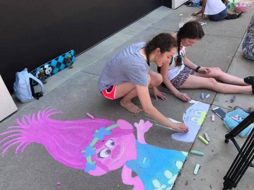Two girls create colorful chalk drawings on the sidewalk, featuring a pink character with a large hairstyle.