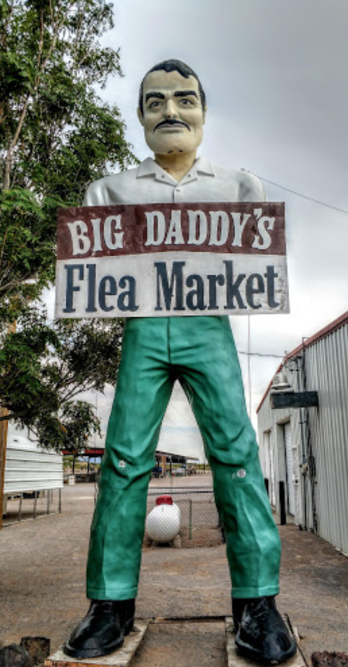 A large statue of a man in green pants and black shoes holding a sign for "Big Daddy's Flea Market."