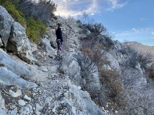 A person in activewear stands on a rocky hiking trail surrounded by sparse vegetation and a clear blue sky.