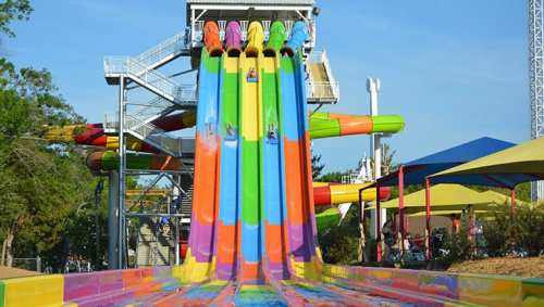 Colorful water slides with people sliding down, surrounded by greenery and a clear blue sky.