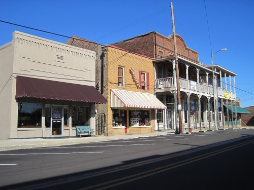 Historic buildings line a quiet street, featuring storefronts with awnings and a clear blue sky above.