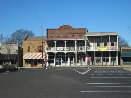 Historic storefronts with balconies and signs, set against a clear blue sky, in a small town. Empty parking lot in front.