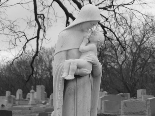 A black and white statue of a mother holding a child, set against a blurred cemetery background.