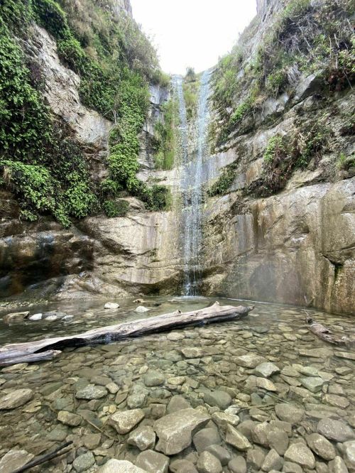 A serene waterfall cascading over rocky cliffs into a clear pool, surrounded by lush greenery and smooth stones.