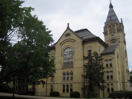 Historic yellow brick building with a tall spire, surrounded by trees under a cloudy sky.