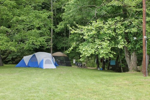 A campsite with a blue and white tent, a screened shelter, and chairs, surrounded by lush green trees.