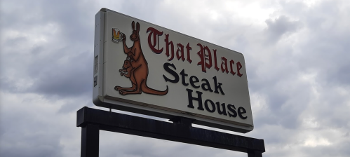 Sign for "That Place Steak House" featuring a kangaroo holding a beer, set against a cloudy sky.