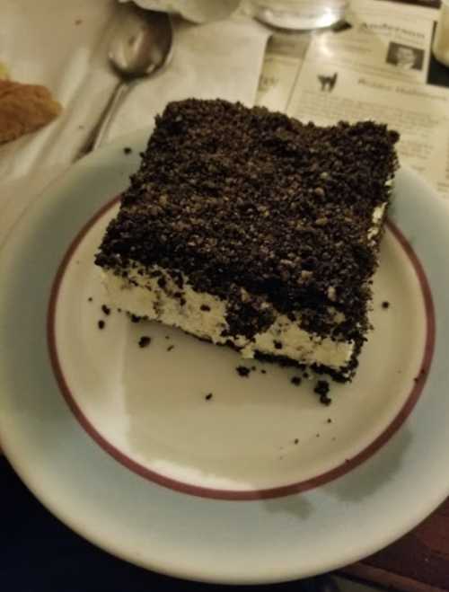 A slice of dessert with layers of cream and crushed chocolate cookies on a white plate.