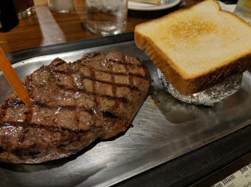 A grilled steak on a metal plate next to a slice of toasted bread wrapped in foil.