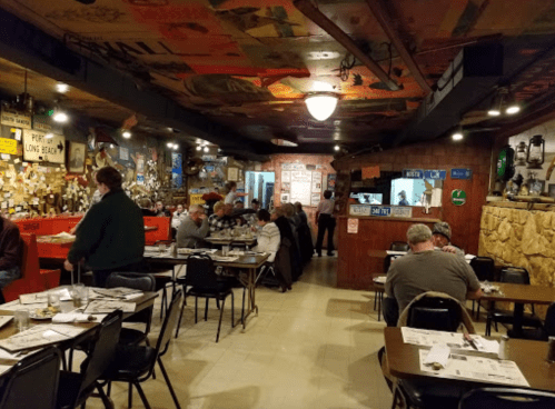 A cozy restaurant interior with wooden decor, diners at tables, and a staff member serving in the background.