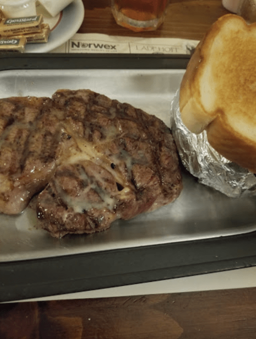 A grilled steak with melted cheese, served on a metal plate alongside a slice of toasted bread.