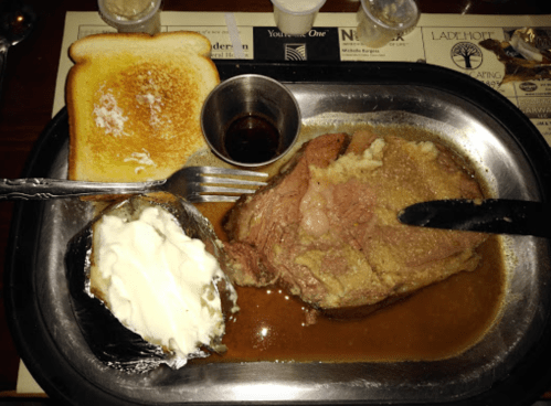 A plate with sliced prime rib, a baked potato with sour cream, toast, and a small cup of gravy.