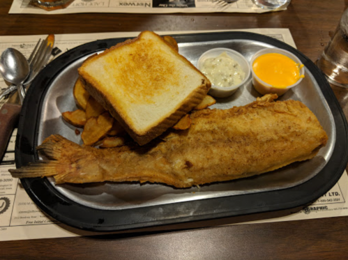 A plate with a large fried fish, toast, and two dipping sauces, served with potato wedges on a wooden table.