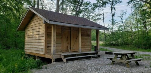 A small wooden cabin with a sloped roof, surrounded by trees, next to a gravel path and a picnic table.