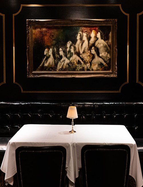 A dimly lit restaurant table with a white tablecloth, a small lamp, and a large painting of women on the wall.