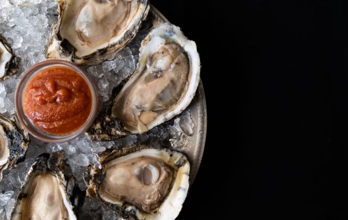 A platter of fresh oysters on ice, accompanied by a small bowl of cocktail sauce.