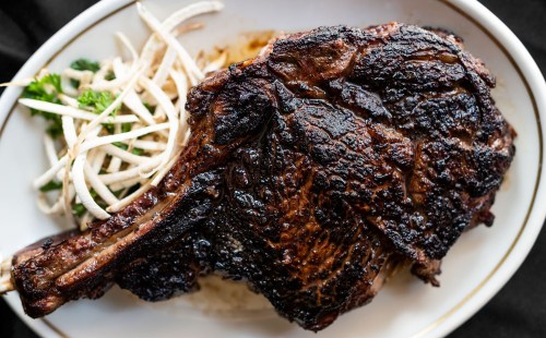 A perfectly cooked, charred ribeye steak served with fresh greens and thinly sliced vegetables on a white plate.