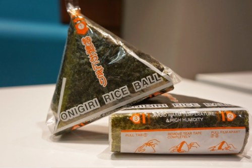 Two packaged onigiri rice balls, one triangular and one rectangular, wrapped in seaweed with instructions on the packaging.