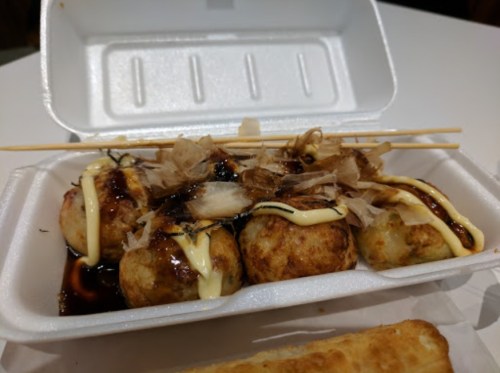 A takeout container with takoyaki balls topped with bonito flakes, drizzled with sauce and mayonnaise.