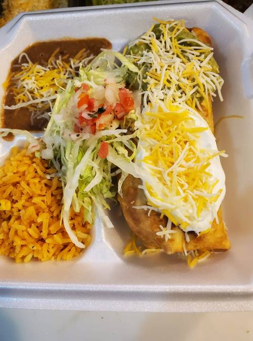 A takeout container with a crispy burrito topped with cheese, sour cream, lettuce, and tomatoes, served with rice and beans.