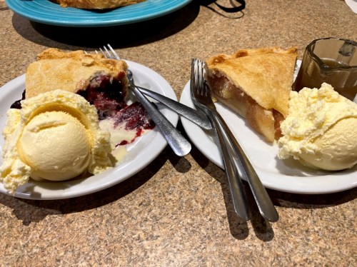 Two plates of pie with scoops of vanilla ice cream, one with berry pie and the other with apple pie, on a table.