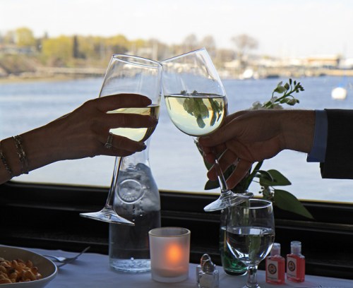 Two hands clink wine glasses filled with white wine, with a scenic waterfront view in the background.