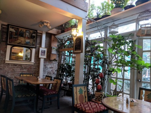 Cozy café interior with wooden tables, plants, and artwork, featuring large windows and a warm, inviting atmosphere.