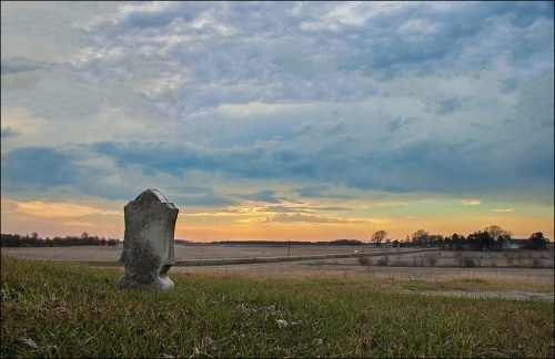 A weathered gravestone stands in a grassy field under a dramatic sky at sunset, with distant trees and open land.