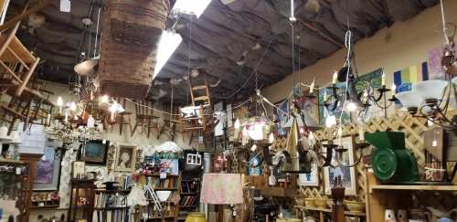 A cluttered antique shop filled with various furniture, chandeliers, and artwork, with chairs hanging from the ceiling.