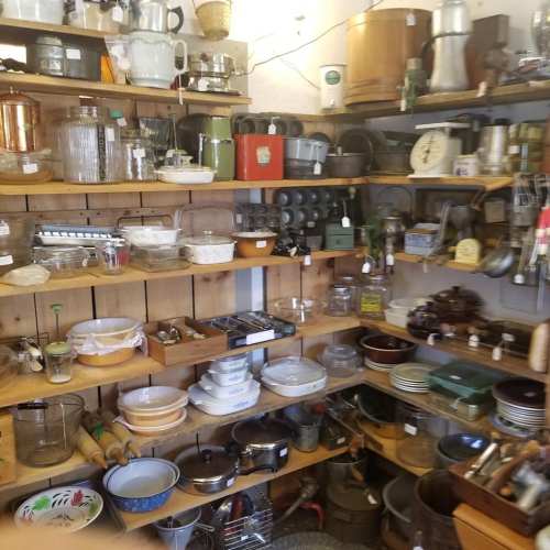 A cluttered display of vintage kitchenware and utensils on wooden shelves, featuring glass, metal, and ceramic items.