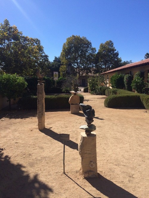 A sunny garden with sculptures, trees, and neatly trimmed hedges under a clear blue sky.
