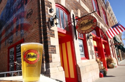 A pint of beer in front of Firehouse Brewing Co. with a brick building and American flag in the background.