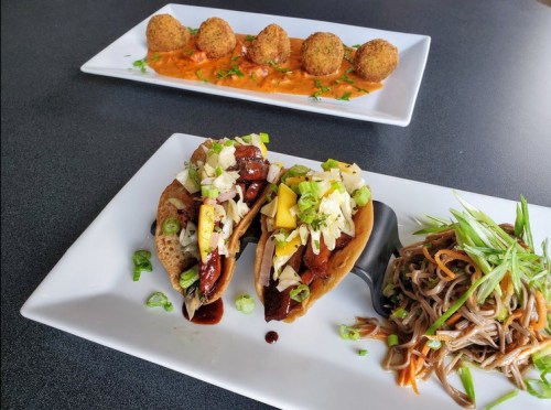 Two tacos with toppings, served alongside a portion of noodles and a plate of croquettes in a creamy sauce.