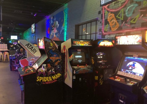 A row of vintage arcade machines, including Spider-Man and Atari, in a colorful, neon-lit gaming venue.
