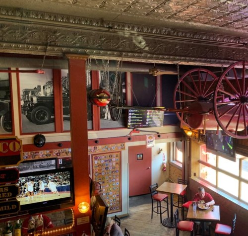A cozy bar interior with vintage decor, including old photos and a wagon wheel, and a TV showing a sports game.