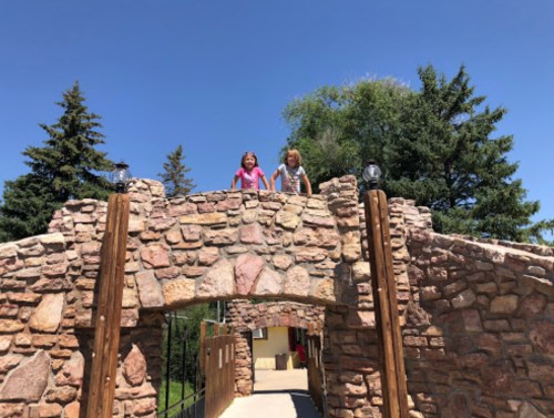 Two children stand on a stone bridge, smiling under a clear blue sky, surrounded by trees.